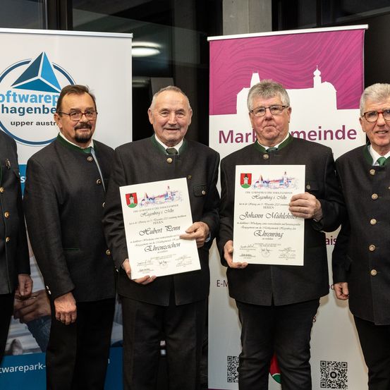 Fünf Männer stehen vor Bannern und lächeln. Zwei halten Urkunden, eine mit 'Hubert Dem' und die andere mit 'Johann Muhlehner'. Sie tragen traditionelle Kleidung mit grünen Krawatten und Medaillen. Das Banner hinter ihnen lautet 'Marktgemeinde'.