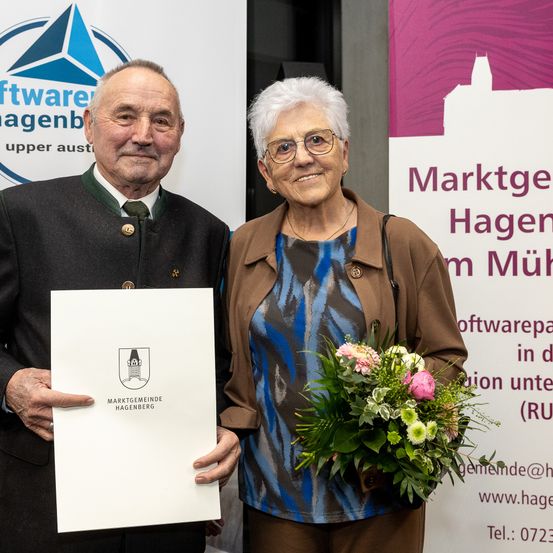 Ein älterer Mann und eine ältere Frau stehen nebeneinander und halten ein Zertifikat und einen Blumenstrauß. Der Mann trägt eine Medaille und die Frau trägt eine Brille. Sie stehen vor einem Banner.