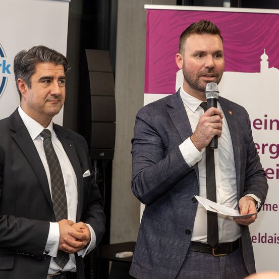 Zwei Männer in Anzügen und Krawatten stehen vor einem Mikrofon und einem Banner. Der Mann auf der rechten Seite hält das Mikrofon und scheint zu sprechen. Der Mann auf der linken Seite schaut zu.