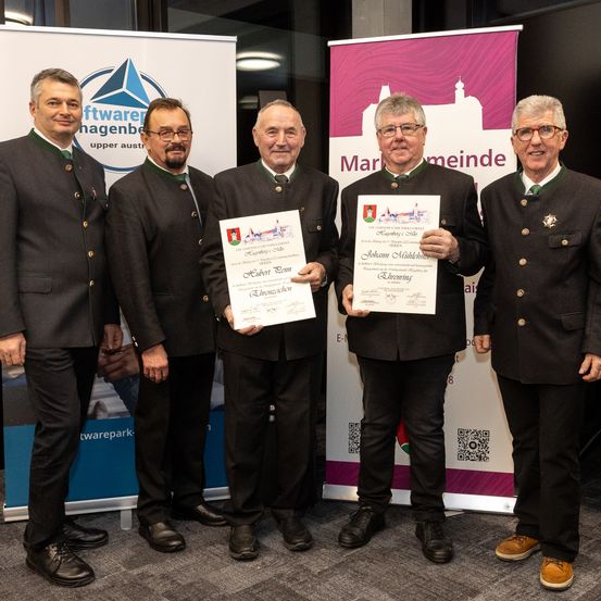 Fünf Männer in traditioneller Kleidung stehen in einer Reihe und halten Urkunden. Der Mann in der Mitte hält eine Urkunde mit dem Namen Johann Mühlreiter. Hinter ihnen sind Banner mit Logos und Text.