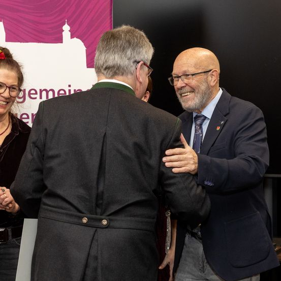 Drei Personen stehen eng beieinander in einer formellen Umgebung. Ein Mann im Anzug lächelt und schüttelt einem anderen Mann die Hand. Hinter ihnen lächelt eine Frau mit Brille. Ein Banner ist im Hintergrund sichtbar.
