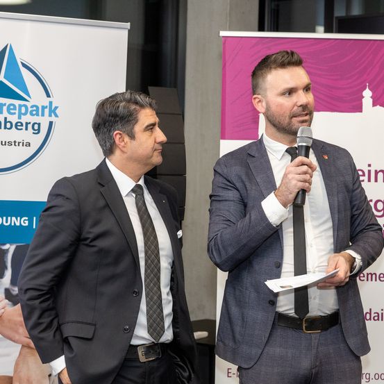 Zwei Männer in Anzügen und Krawatten, einer spricht in ein Mikrofon, der andere hört zu. Hinter ihnen sind Banner mit Logos und Text zu sehen.