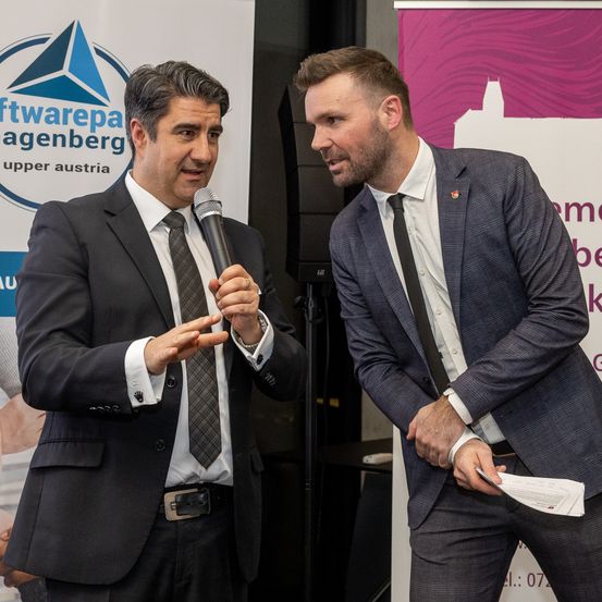 Zwei Männer in Anzügen sprechen bei einer Konferenz miteinander. Einer hält ein Mikrofon und der andere ein Blatt Papier. Sie stehen vor einem Banner mit einem Logo.