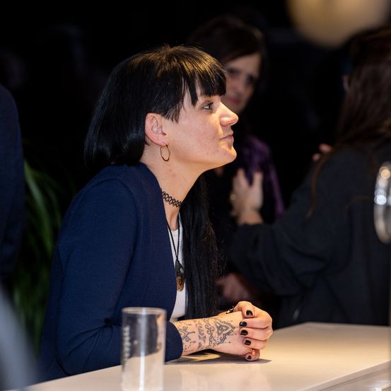 Eine Frau mit schwarzen Haaren und Tattoos sitzt an einem Tisch und schaut nachdenklich zur Seite. Ein Glas Wasser steht vor ihr. Andere Personen sind anwesend, aber unscharf.