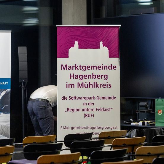 Ein Banner für die Marktgemeinde Hagenberg im Mühlkreis, die Softwarepark-Gemeinschaft in der Region Untere Feldaist (RUF). Eine Person ist dabei zu sehen, wie sie Stühle in einem Konferenzraum anordnet.