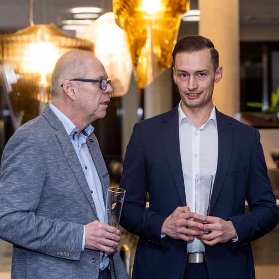 Zwei Männer stehen in einem gut beleuchteten Raum und halten beide Glaspreise in den Händen. Der Mann auf der linken Seite trägt einen grauen Anzug, und der Mann auf der rechten Seite trägt einen dunkelblauen Anzug. Hinter ihnen hängen dekorative Kronleuchter von der Decke.