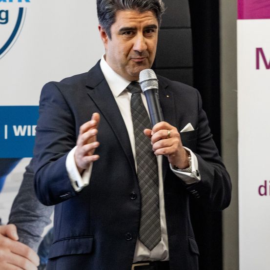 Ein Mann in einem schwarzen Anzug und Krawatte spricht in ein Mikrofon bei einer Veranstaltung. Hinter ihm ist eine weiße Kulisse mit Text und Logos zu sehen.