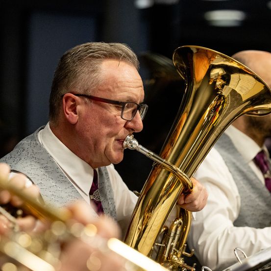 Ein Musiker mit Brille und Weste spielt während eines Konzerts eine Tuba. Ein weiterer Musiker ist im Hintergrund zu sehen.