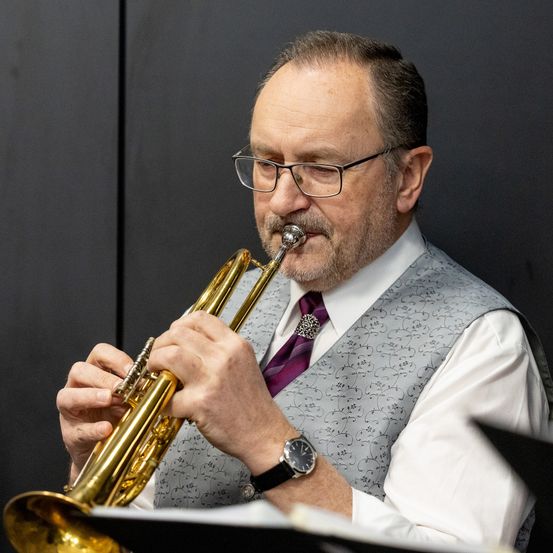 Ein mittelalte Mann mit Brille und Uhr spielt Trompete. Er ist in einem weißen Hemd und grauer Weste gekleidet.
