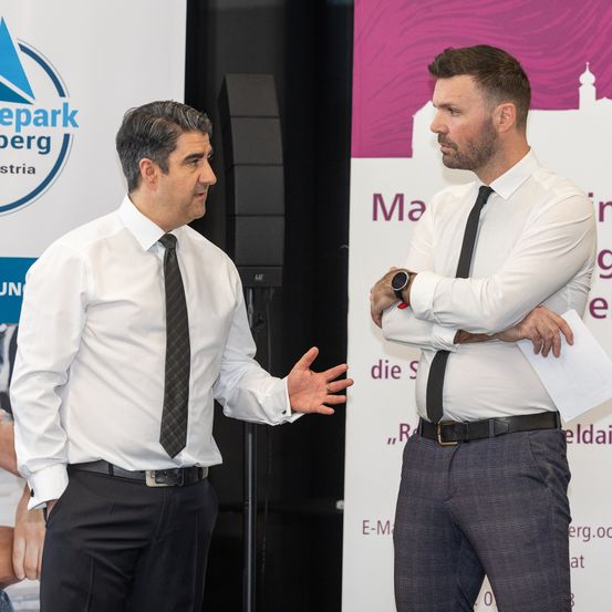 Zwei Männer in formeller Kleidung stehen und unterhalten sich bei einer formellen Veranstaltung, einer gestikuliert, während der andere aufmerksam zuhört. Hinter ihnen befindet sich ein Banner mit Logo und Text.