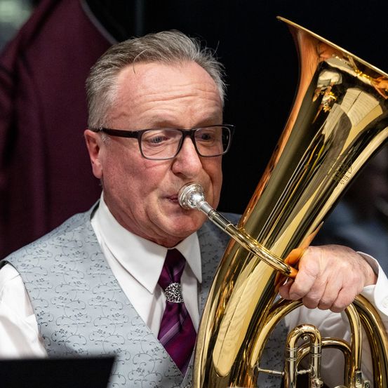 Ein älterer Mann mit Brille spielt während einer Aufführung eine Tuba. Er trägt ein weißes Hemd, eine gemusterte Weste und eine lila Krawatte.