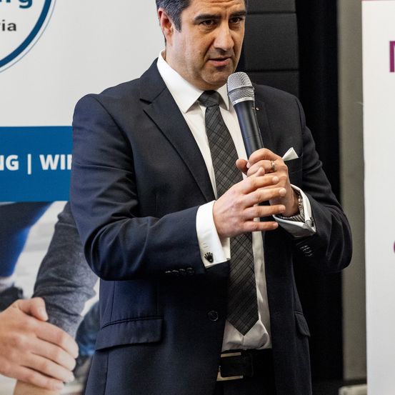 Ein Mann in einem schwarzen Anzug hält ein Mikrofon und scheint bei einer Veranstaltung zu sprechen. Der Hintergrund zeigt ein blaues Banner mit weißem Text und Logos.