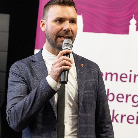 Ein Mann in einem Anzug hält ein Mikrofon und spricht vor einer Kulisse mit Stadtsilhouette und Text auf Deutsch.
