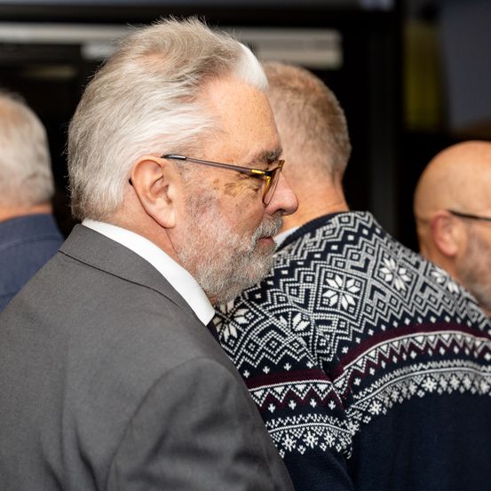 Ein Mann mit weißem Haar und Bart trägt eine Brille und steht in einer Gruppe von Männern, von denen zwei gemusterte Pullover tragen.
