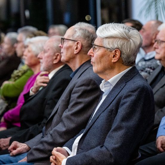 Eine Gruppe älterer Menschen sitzt in einer Reihe, wahrscheinlich in einem Konferenzsaal. Der Mann vorne trägt eine Brille und sieht nach vorne.
