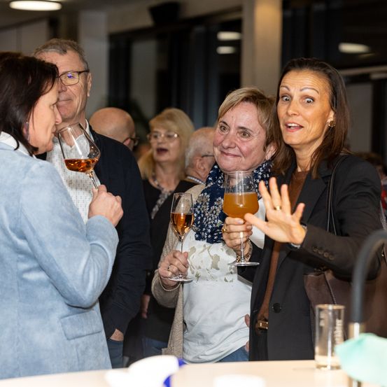 Mehrere Erwachsene sind drinnen bei einer Zusammenkunft, wobei drei Frauen lächelnd Weingläser halten. Ein Mann hinter ihnen scheint mit einer anderen Frau zu sprechen. Vor ihnen steht ein Tisch mit Weingläsern.