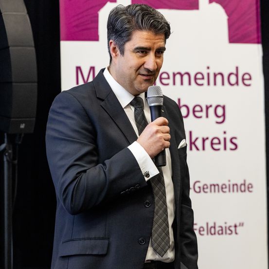 Ein Mann in einem Anzug spricht in ein Mikrofon vor einem Banner, das 'Meine Gemeinde' und 'Feldais' besagt.