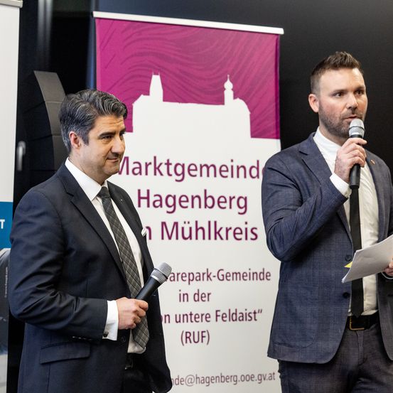 Zwei Männer in Anzügen sprechen in Mikrofone vor einem Banner für die Marktgemeinde Hagenberg im Mühlkreis.