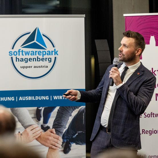 Ein Mann hält eine Rede vor einem Softwarepark Hagenberg-Schild. Er hält ein Mikrofon, und die Menschen im Publikum haben ihre Hände zusammengepresst.