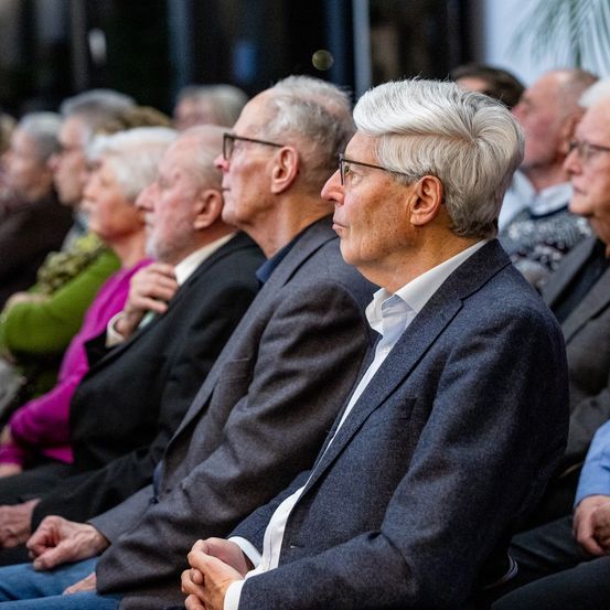 Eine Gruppe älterer Menschen sitzt in einem Auditorium und trägt formelle Kleidung. Der Mann in der vordersten Reihe trägt eine Brille und hat die Hände ineinander gefaltet.