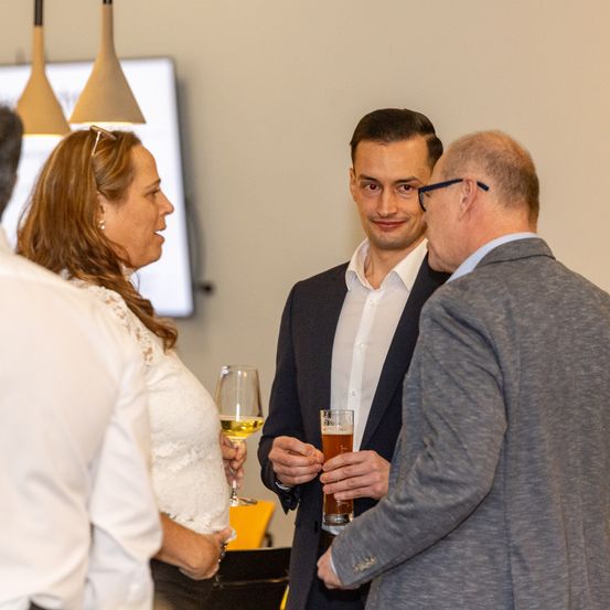 Drei Erwachsene unterhalten sich in einem Innenbereich. Ein Mann hält ein Glas Bier, während eine Frau ein Glas Wein hält.
