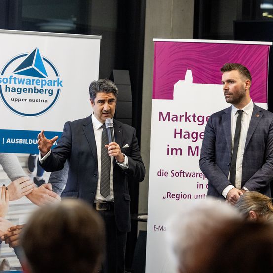 Zwei Männer in Anzügen stehen neben Bannern. Ein Mann spricht in ein Mikrofon, während der andere zuhört. Sie befinden sich auf einer Konferenz.