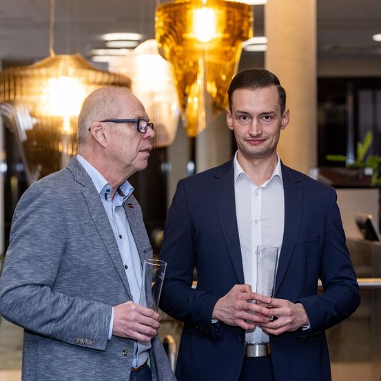 Zwei Männer stehen nebeneinander und halten Trophäen. Sie lächeln und scheinen miteinander zu sprechen. Sie tragen beide formelle Anzüge. Hinter ihnen hängt eine leuchtend gelbe Kronleuchter.