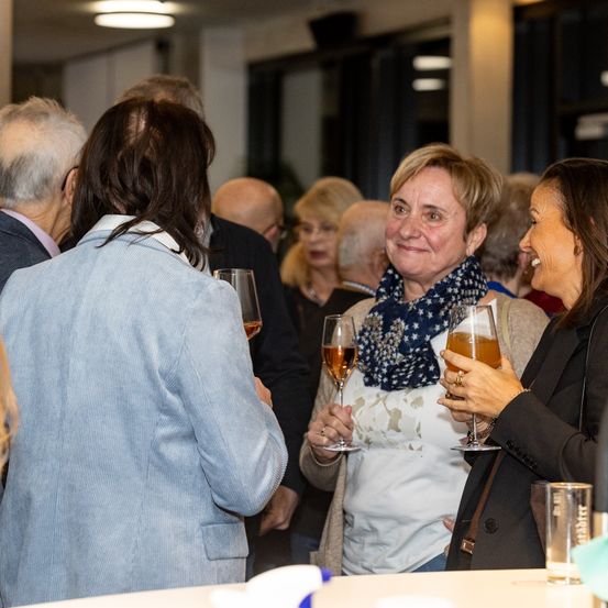 Eine Gruppe von Menschen, die bei einer Veranstaltung stehen und lächeln, während sie Weingläser halten. Zwei Frauen in der Mitte lachen.