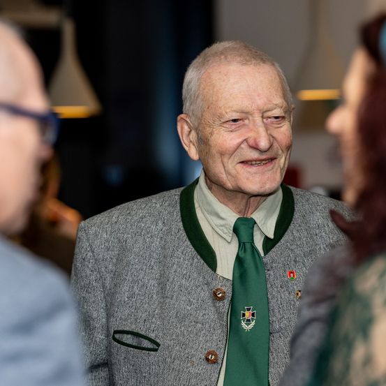 Ein älterer Mann mit einer grünen Krawatte mit weißem Emblem und einem grauen Jackett mit grünen Kragen lächelt und scheint mit einer Frau zu sprechen.