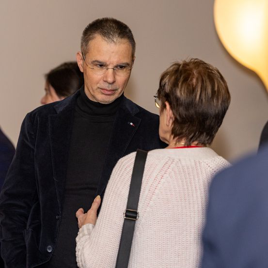 Ein Mann mit Brille und schwarzem Rollkragen spricht mit einer Frau mit kurzen Haaren und einem weißen Pullover. Sie sind drinnen mit einer gelben Lampe in der Nähe.