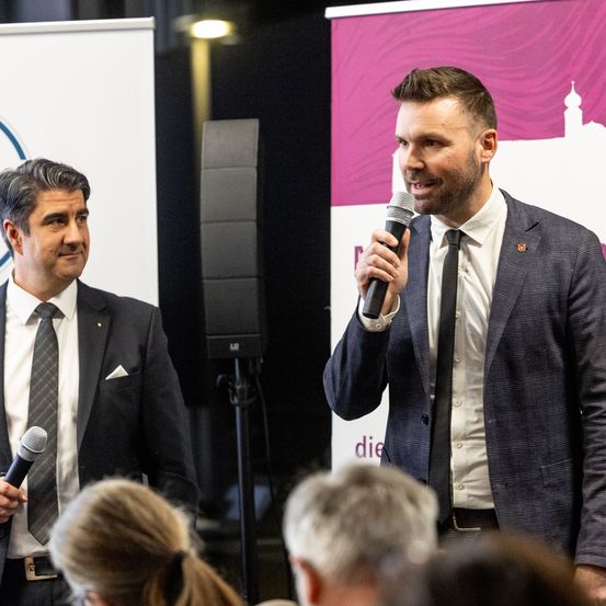 Zwei Männer in Anzügen stehen vor einem Mikrofon und einem Banner. Ein Mann hält das Mikrofon und lächelt, während der andere neben ihm steht. Dahinter steht ein Lautsprecher auf einem Ständer.
