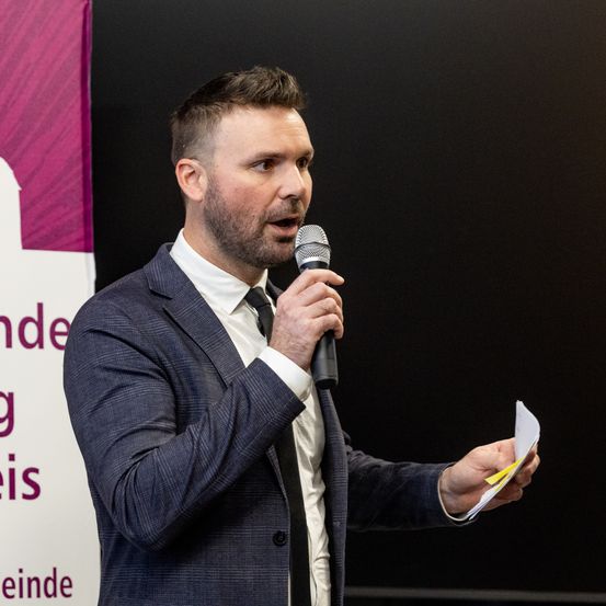 Ein Mann im Anzug spricht in ein Mikrofon vor einem dunklen Hintergrund mit einem Banner.