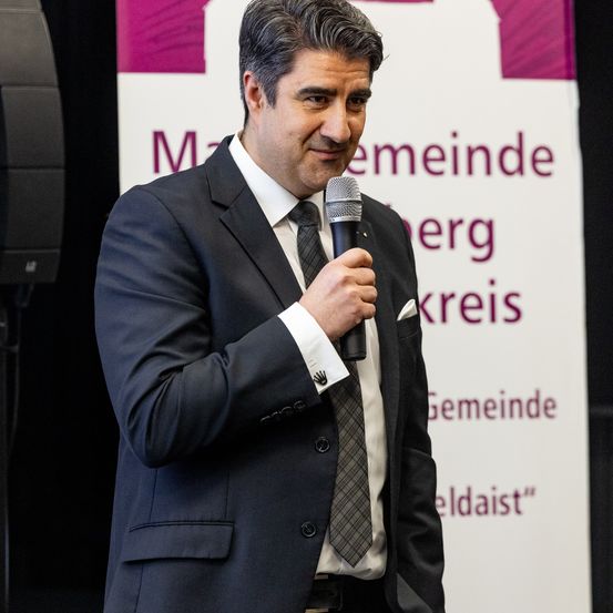 Ein Mann in Anzug und Krawatte spricht in ein Mikrofon vor einem Schild.