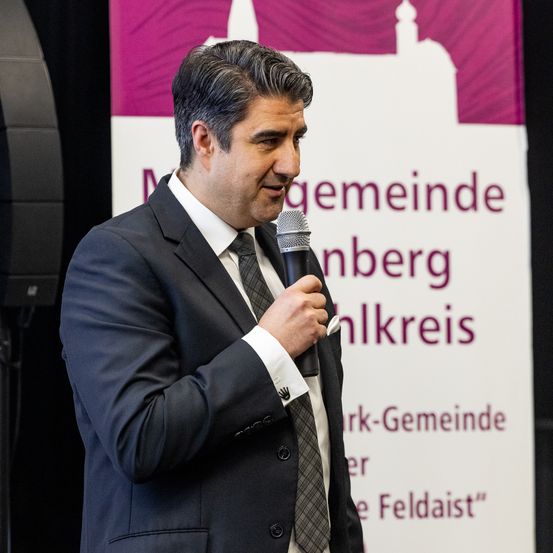 Ein Mann in Anzug und Krawatte steht vor einem Mikrofon und spricht bei einer Veranstaltung. Ein lila-weißes Banner hinter ihm trägt die Aufschrift 'Gemeindenburg' und 'Landkreis'.