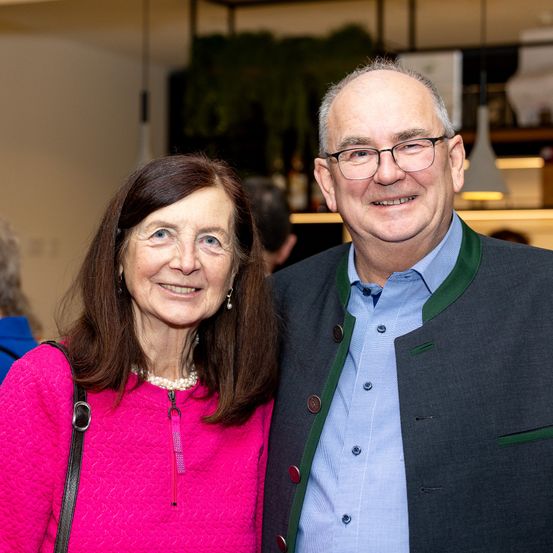 Ein älteres Paar steht eng beieinander und lächelt für ein Foto. Die Frau trägt eine rosa Jacke und eine Perlenkette, während der Mann ein blaues Hemd und einen grauen Anzug mit grünen Akzenten trägt. Hinter ihnen zeigt ein verschwommener Hintergrund einen Raum mit Regalen und einer hängenden Lampe.
