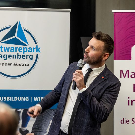 Ein Mann spricht in ein Mikrofon vor einem Softwarepark Hagenberg Banner, mit einem teilweise sichtbaren Banner im Hintergrund, das 'Markt' steht.