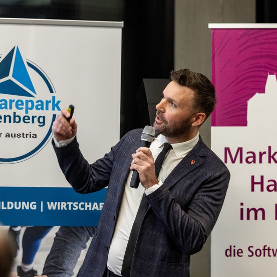 Ein Mann im Anzug, der ein Mikrofon hält, steht vor einem Schild mit Logo und Text.