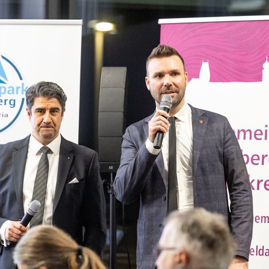 Zwei Männer stehen an einem Podium, einer hält ein Mikrofon. Ein Mann spricht, während der andere zuhört. Hinter ihnen ein Banner mit einem abstrakten Design.