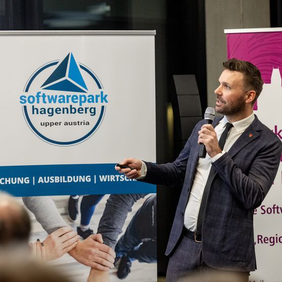Ein Mann in einem Anzug spricht in ein Mikrofon vor einem Softwarepark Hagenberg Banner, mit Händen die Teamarbeit im Hintergrund symbolisieren.