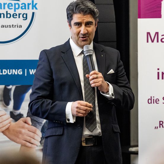 Ein Mann in einem Anzug hält ein Mikrofon und spricht vermutlich bei einer Veranstaltung. Hinter ihm steht ein Schild mit 'Carepark Amberg, Austria'.