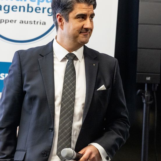 Ein Mann in einem schwarzen Anzug, weißem Hemd und Krawatte lächelt, während er ein Mikrofon hält, möglicherweise bei einer Veranstaltung. Hinter ihm ist ein Schild mit 'Oberösterreich' zu sehen.
