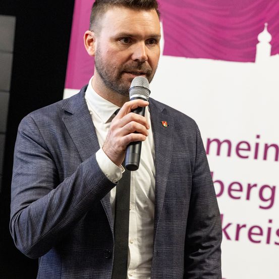Ein Mann in einem grauen Anzug und einer Krawatte steht vor einem Mikrofon und spricht zu einem Publikum. Ein Banner hinter ihm zeigt ein Gebäude mit einer Kuppel und deutschen Text.