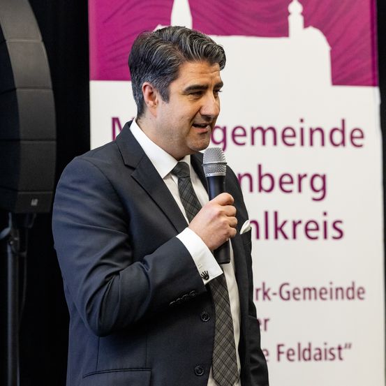 Ein Mann in Anzug und Krawatte hält ein Mikrofon und lächelt bei einer Veranstaltung in der Stadt Nürnberg.