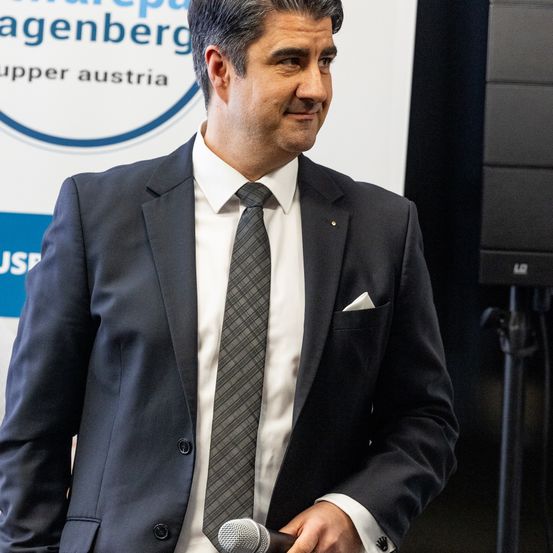 Ein Mann in einem dunklen Anzug und Krawatte, lächelnd und ein Mikrofon haltend, steht vor einem Banner mit Text.
