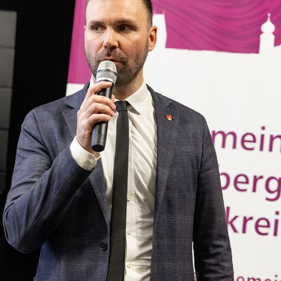Ein Mann im Anzug hält ein Mikrofon in der Hand und hält vermutlich eine Rede bei einer Veranstaltung. Er hat einen ernsten Gesichtsausdruck. Hinter ihm ist ein rosafarbener Banner mit weißer Schrift.