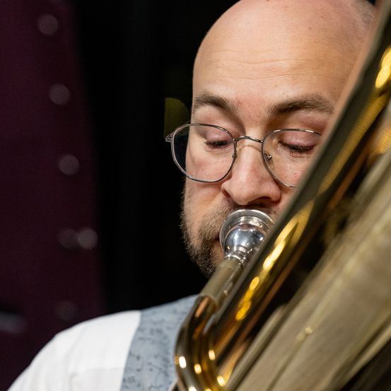 Ein Mann mit Brille spielt ein Blechblasinstrument. Er hat die Augen geschlossen und konzentriert sich auf seine Darbietung.