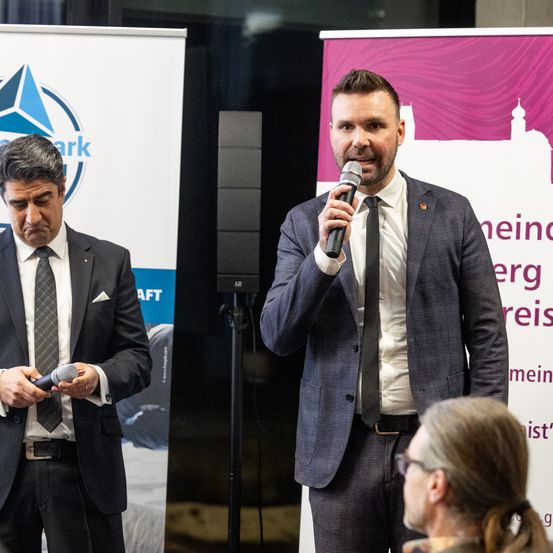 Zwei Männer stehen an einem Rednerpult; einer spricht in ein Mikrofon, während der andere wegschaut. Ein Banner mit 'Meindorf' Text und einem Lautsprecher steht hinter ihnen.