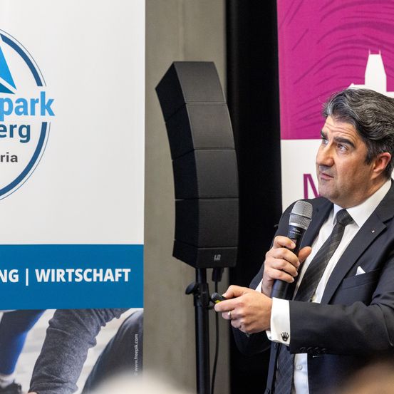 Ein Mann spricht in ein Mikrofon bei einer Veranstaltung. Ein großer Lautsprecher steht hinter ihm, und ein Logo für einen Park befindet sich auf einem nahen Banner.