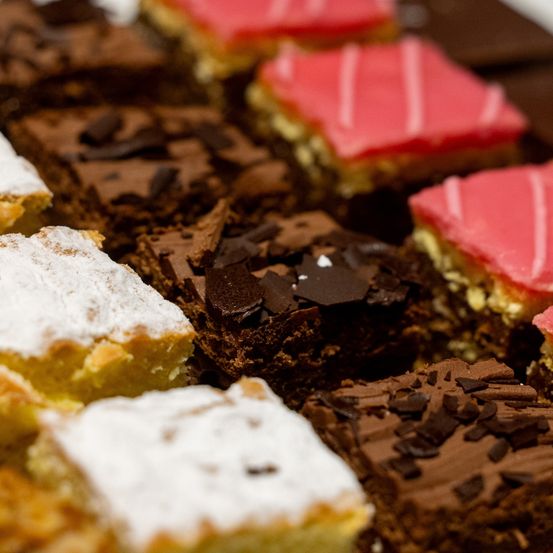 Eine Nahaufnahme verschiedener Desserts auf einem Holztablett, darunter Brownies, weiß pulverisierte Kuchen und ein rot glasierter quadratischer Kuchen.