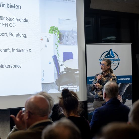 Ein Redner präsentiert vor einer Projektionsleinwand und Publikum. Die Leinwand zeigt Dienstleistungen für Studenten, Beratung und mehr.